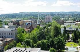 Cityscape with buildings, trees, and a church steeple against a backdrop of wooded hills.