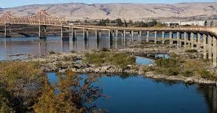 Bridge over water, rocky foreground, mountains in the background, blue sky.