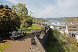 Park pathway overlooking a river with a bench, trees, and buildings under a blue sky.
