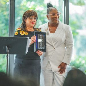 Two women are standing next to each other in front of a window holding a plaque.