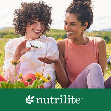 Two women are sitting in a field holding bottles of vitamins.