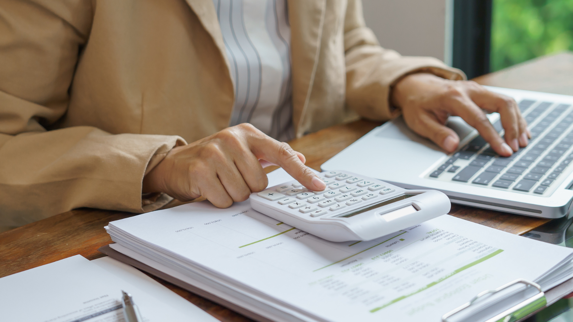 A person in a beige jacket using a calculator while working on a laptop, with tax-related documents and a pen on the desk, focusing on financial calculations for tax purposes.