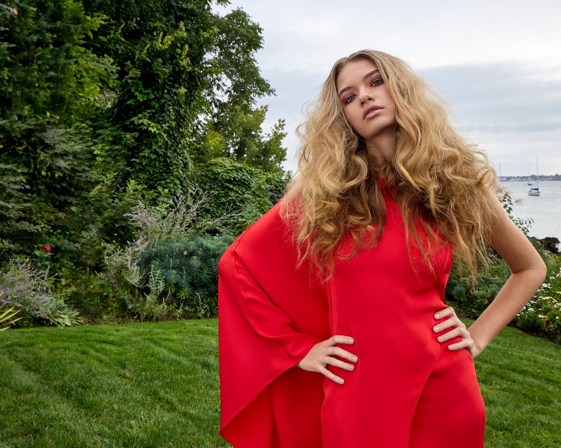 International vogue model in a red dress is standing in the grass with her hands on her hips.