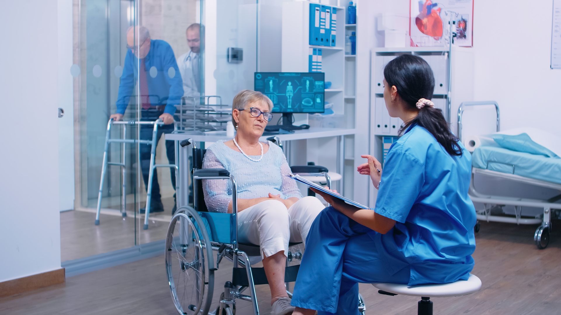 A healthcare worker consults a patient in a wheelchair, with another patient using a walker visible in the background.