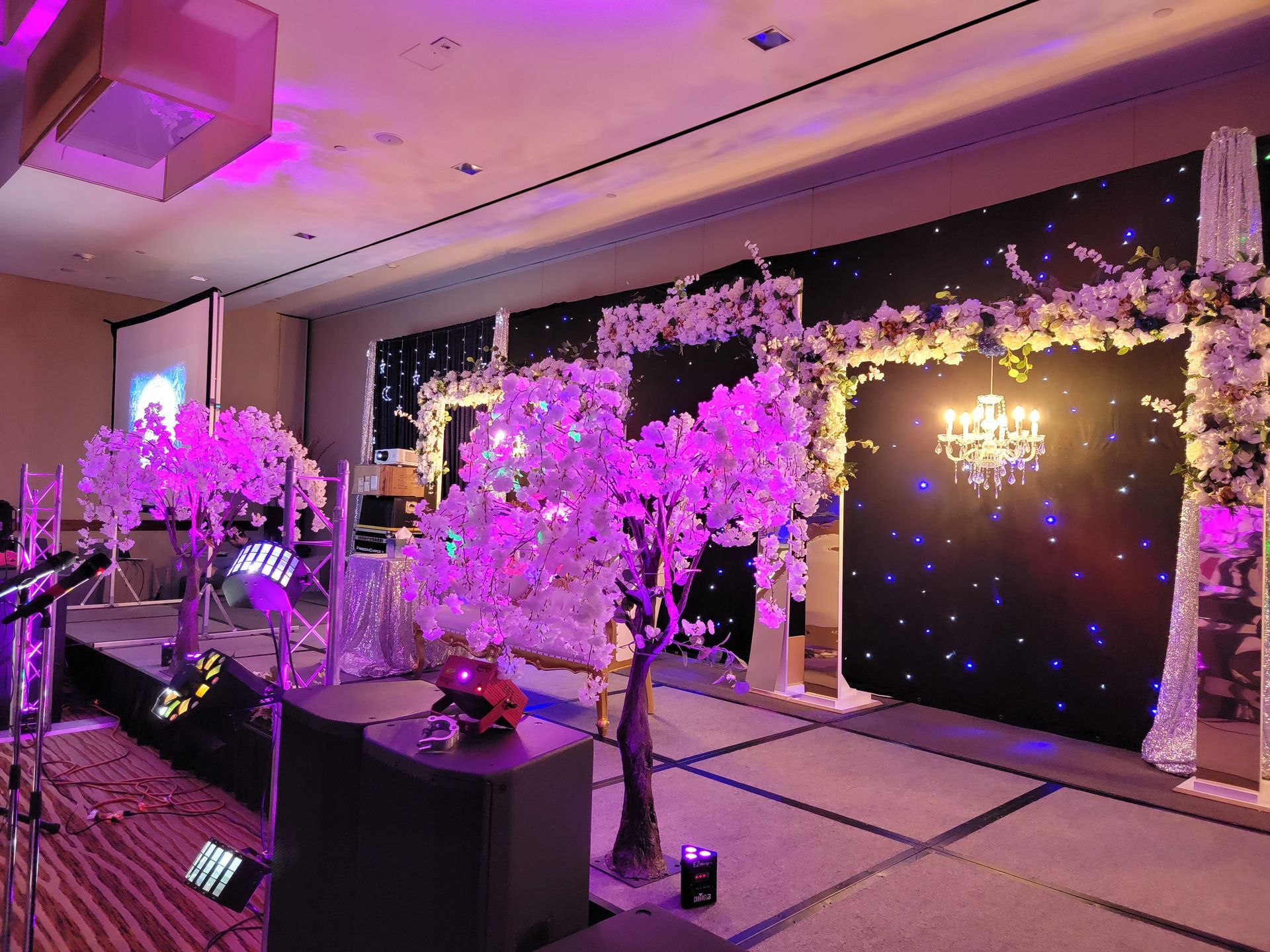 A dimly lit event space featuring a long bar, tables with red tablecloths, seating areas, and hanging globe lighting.