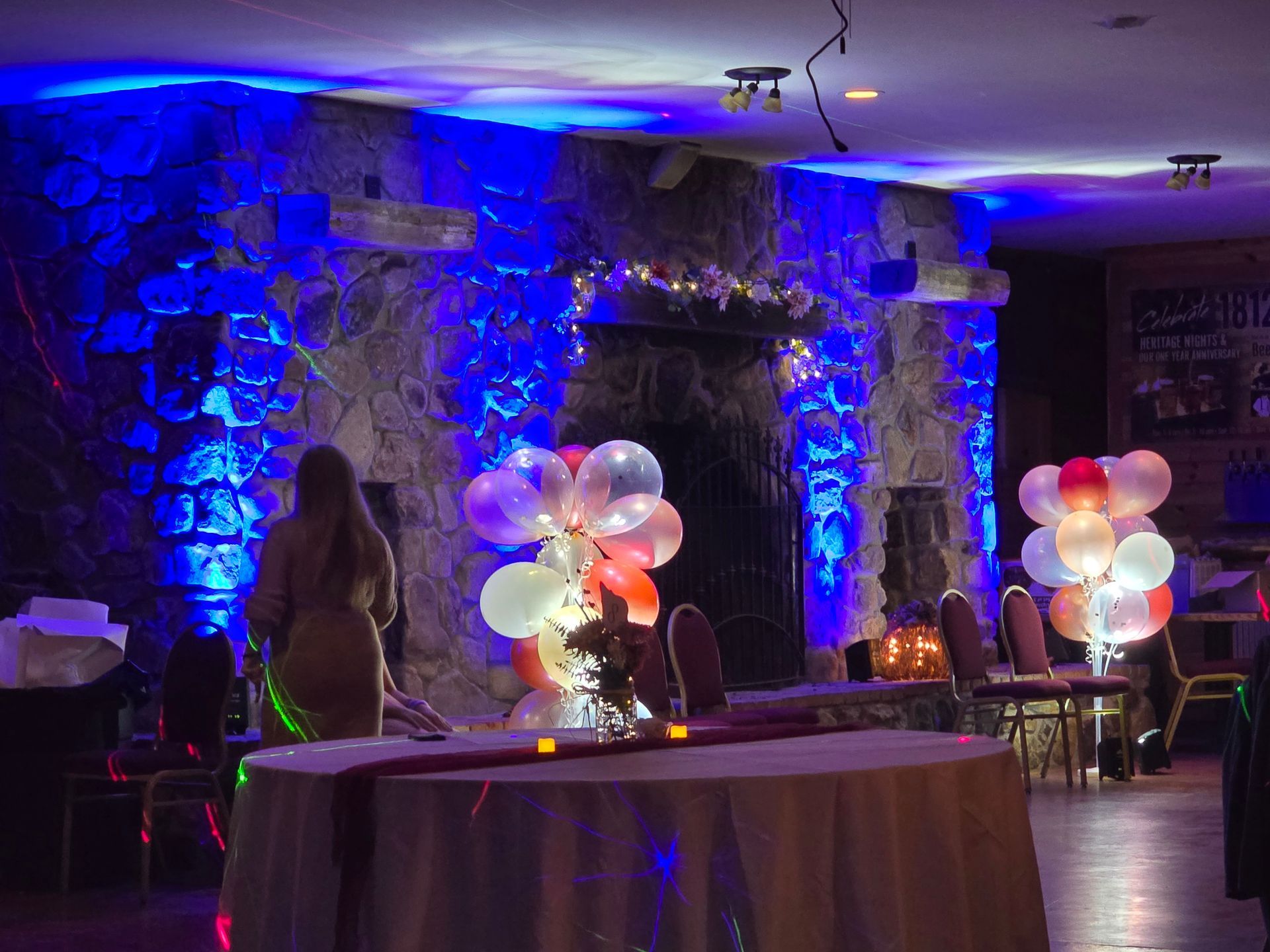 A person in a room with blue ambient lighting stands near a stone fireplace decorated with glowing, illuminated balloons.