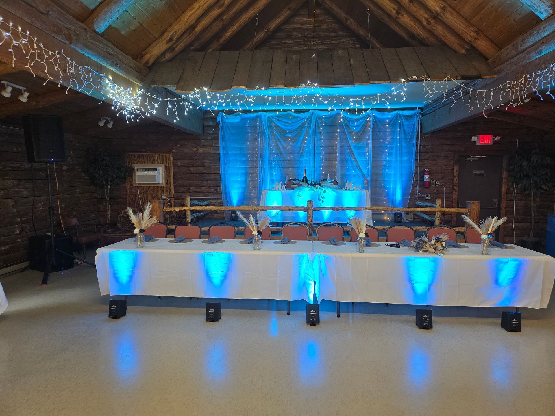 A long wedding head table decorated with white linens and blue uplighting inside a rustic wood-paneled hall.