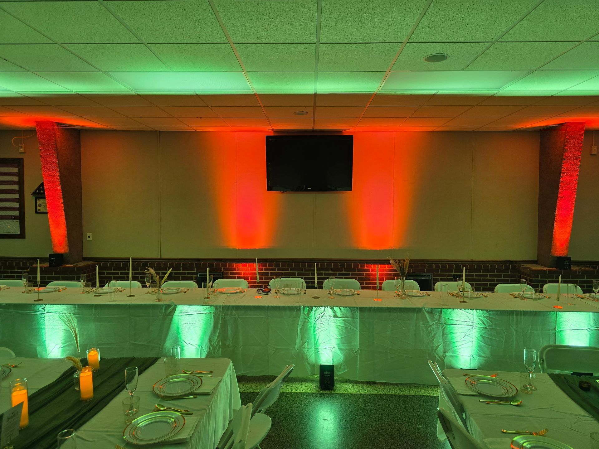 An event space featuring a long table with white tablecloths, decorated with vibrant green and orange up-lighting.