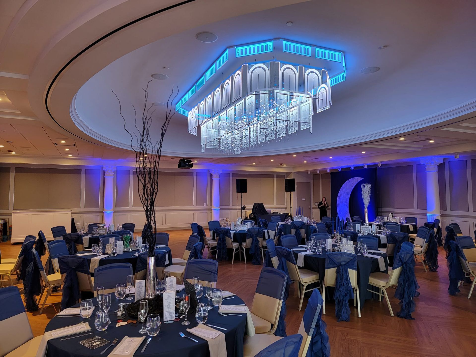 An elegant banquet hall with round tables, navy blue and tan linens, and a large, blue-lit geometric chandelier.