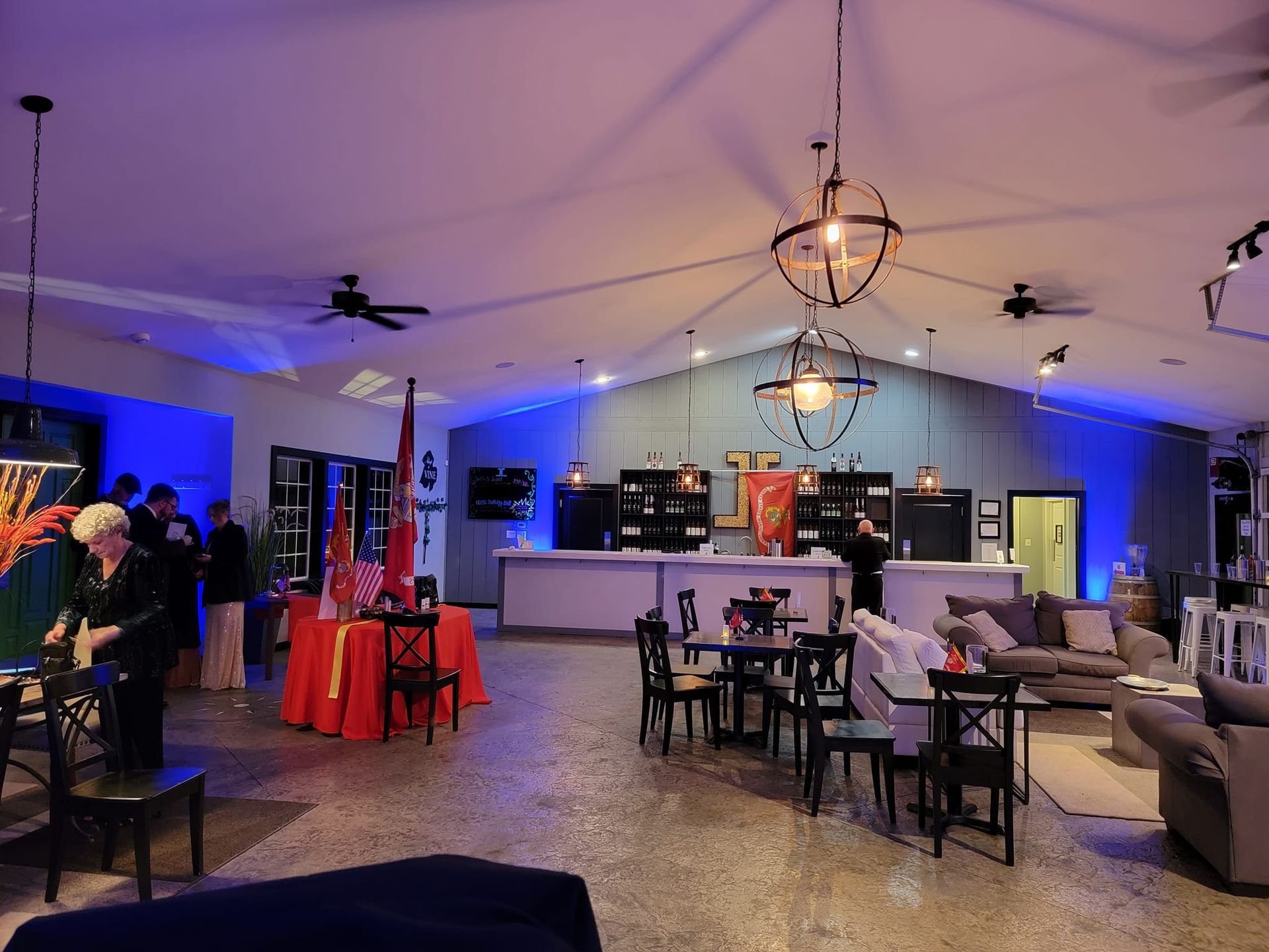 A dimly lit event space featuring a long bar, tables with red tablecloths, seating areas, and hanging globe lighting.