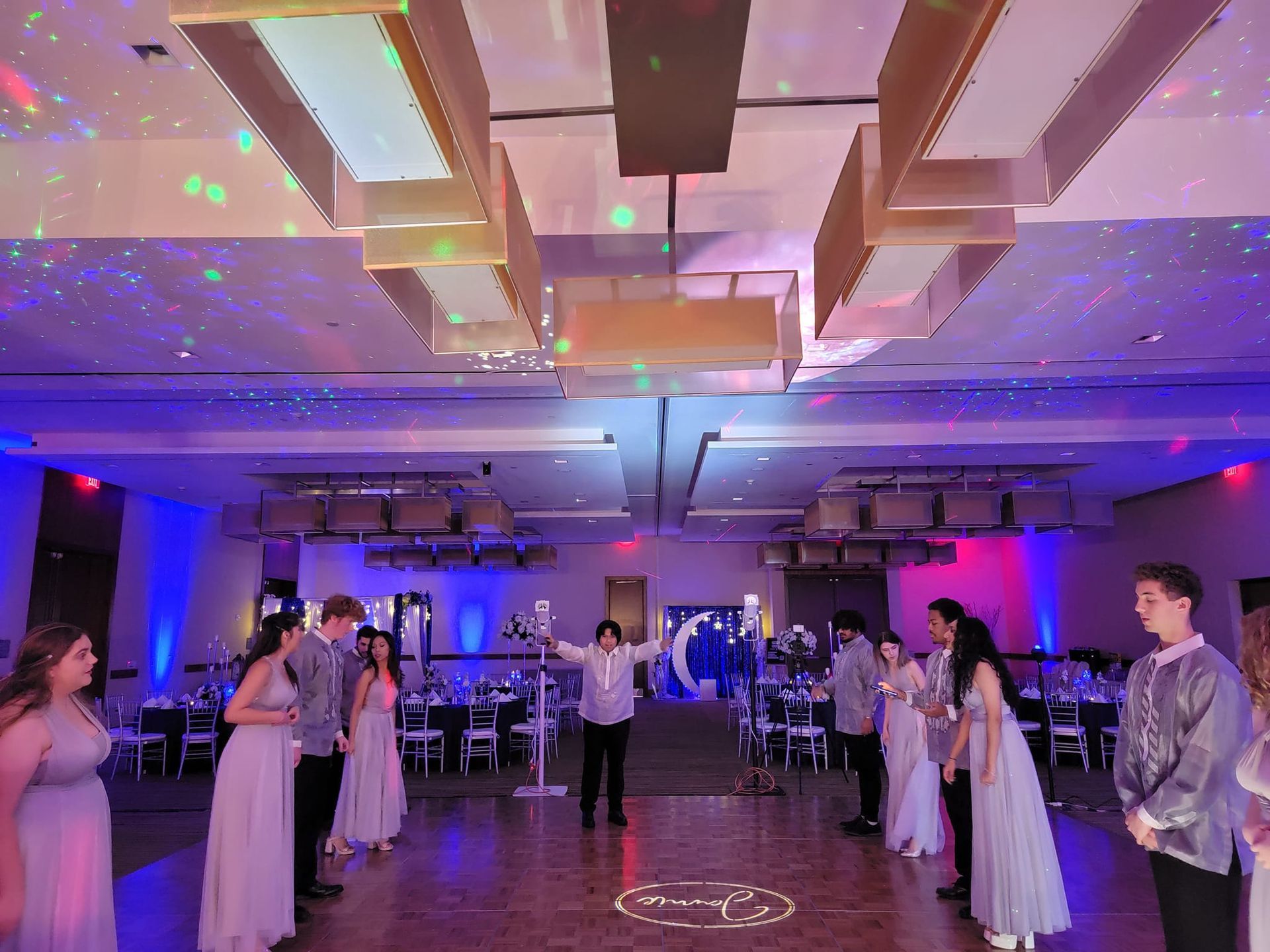 A group in formal wear gathers on a dance floor in a ballroom decorated with purple lighting and disco light effects.