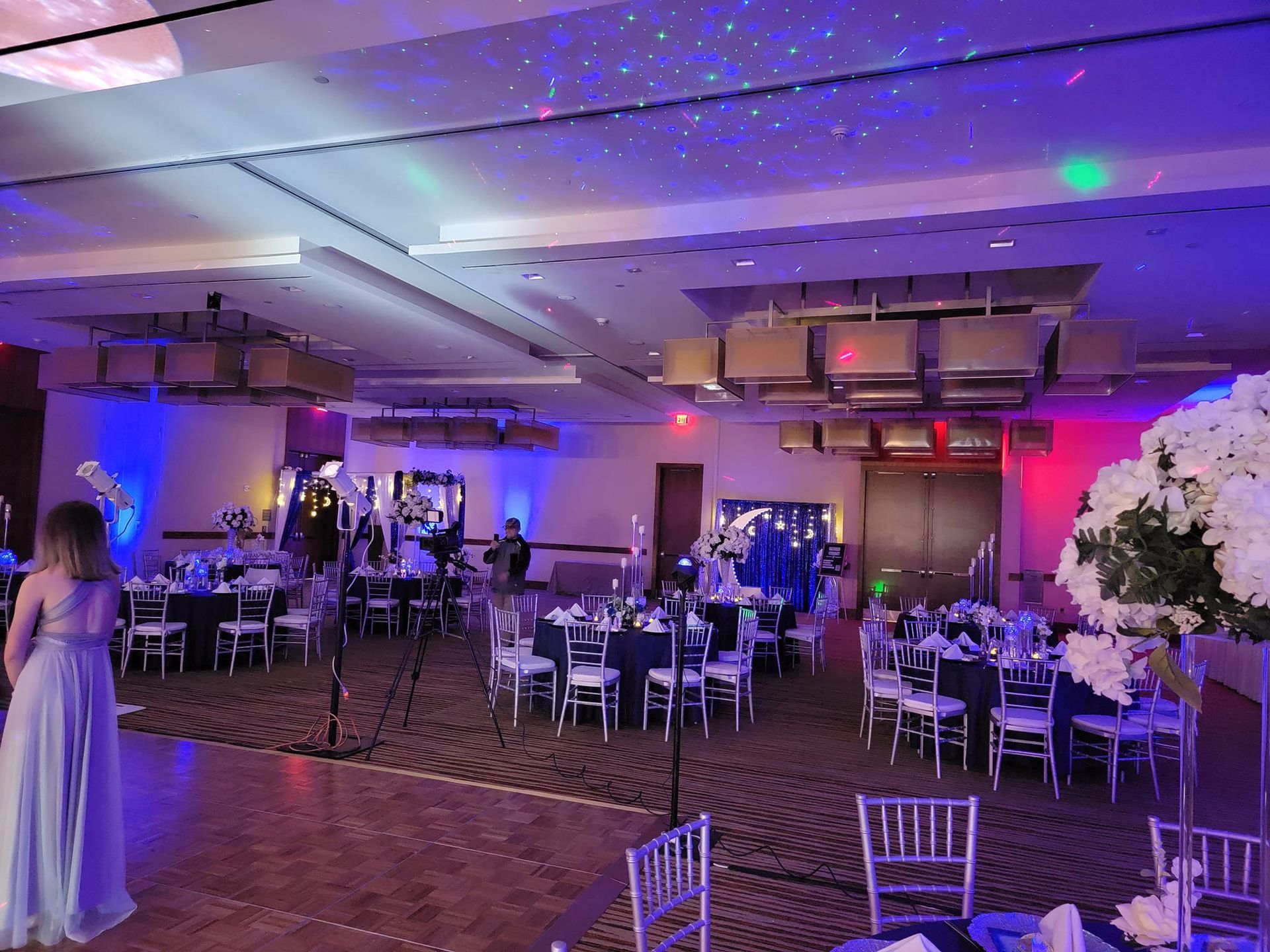 A ballroom set for an event with round tables, white floral centerpieces, purple lighting, and a dance floor.