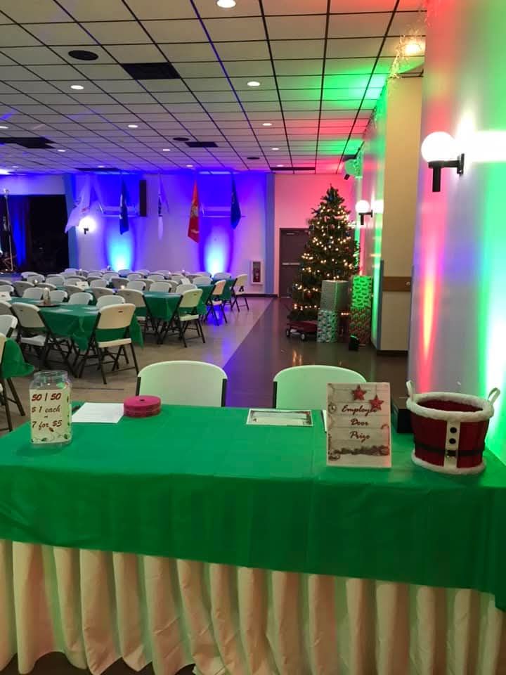 A green-clothed welcome table at a holiday party with a Christmas tree, colorful stage lighting, and chairs in a hall.