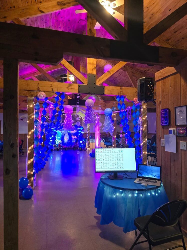 A wooden event hall decorated with blue and purple lighting and balloon garlands, featuring a desk with a computer setup.