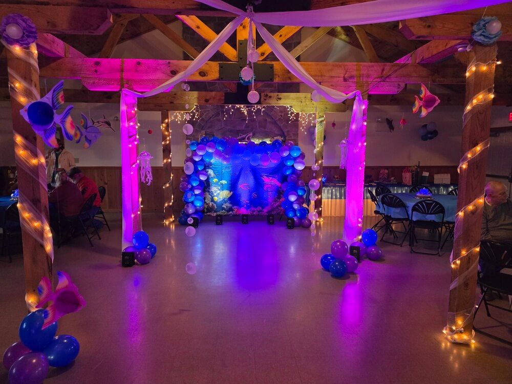 An indoor space decorated for an under-the-sea party with blue and purple balloon arches, fish cutouts, and fairy lights.