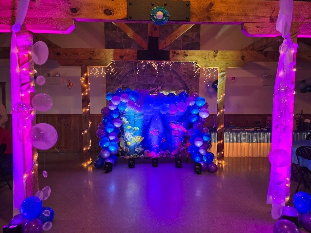 A party room featuring a blue and purple balloon archway against an ocean-themed backdrop, framed by glowing pink columns.