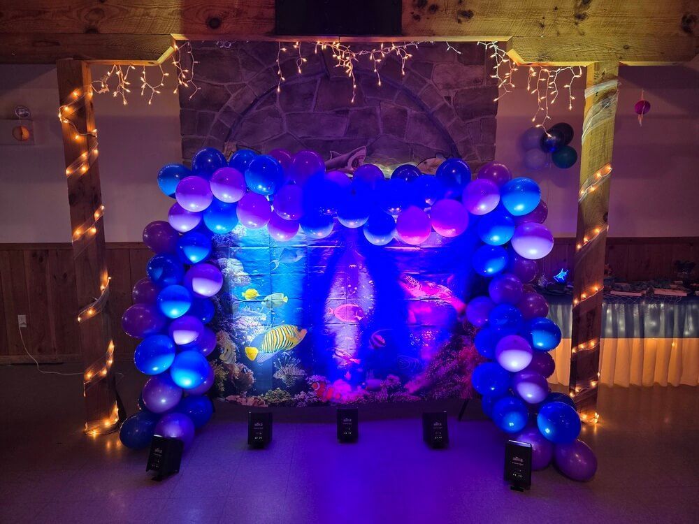 A balloon arch frames an under-the-sea backdrop with string lights on wooden pillars in a dimly lit room.