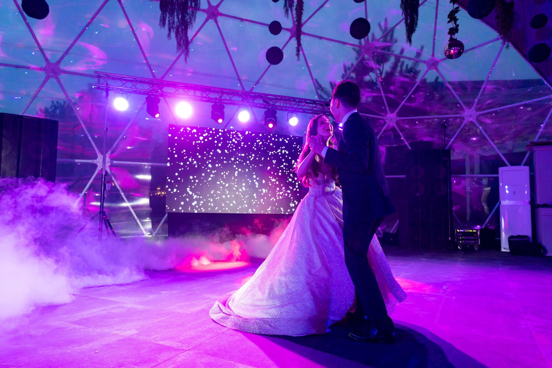A couple dances in a glowing, purple-lit dome structure filled with floor-level fog under a geometric canopy.