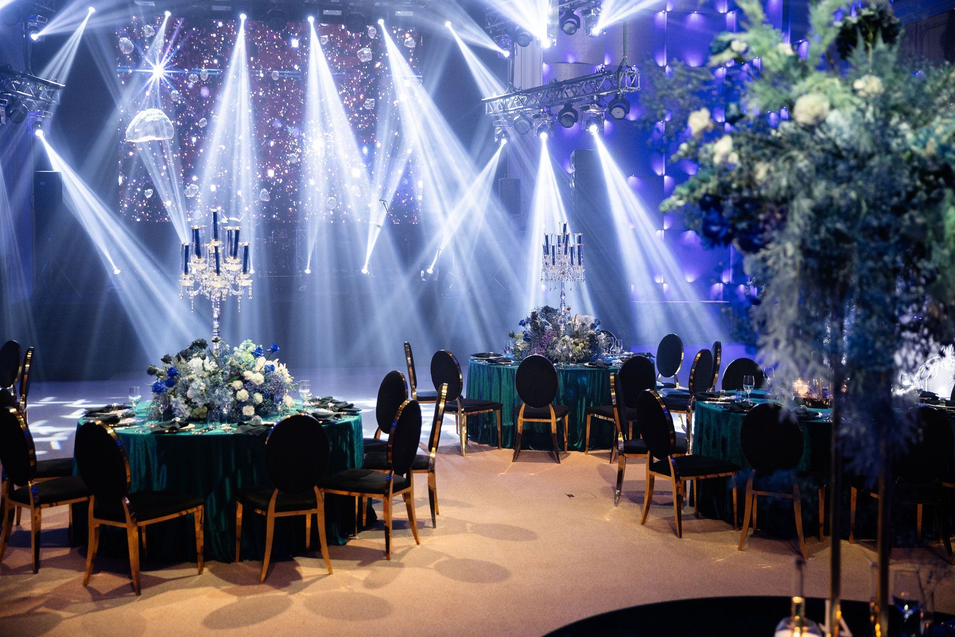 An elegantly decorated event space with round tables, dark teal tablecloths, tall candelabras, and dramatic spotlights.