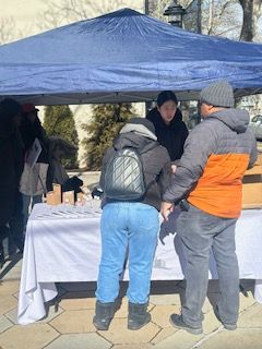 People at a table under a blue tent; one person is helping customers. Outdoors, possibly a market or event.