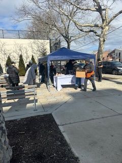 People at a blue tent, possibly an outdoor market. Boxes, table with items, and trees in background.