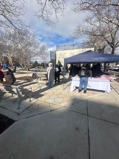 People at a table under a canopy in a park setting. Sunlight and blue sky.