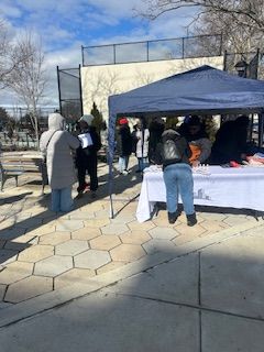 People at a table under a blue tent outdoors, possibly an event or market, on a sunny day.