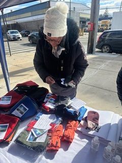 Woman at outdoor table with hats and gloves, looking at an item. Sunny day.