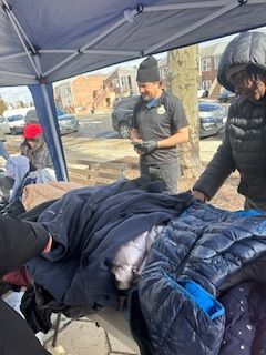 People browsing jackets at an outdoor donation table. A man in a black shirt smiles, others reach for clothing.