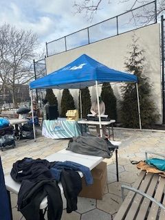 Blue canopy over a table with clothing, a person, and a backdrop of a fence and trees.