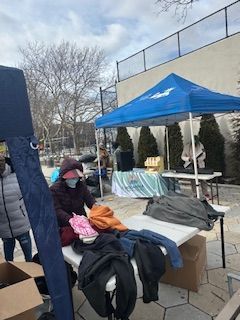People at a table under a blue tent with clothes, boxes, and a speaker outside on a sunny day.