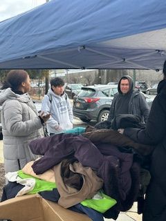 People at table selecting clothes, under a blue canopy. Cars parked behind.