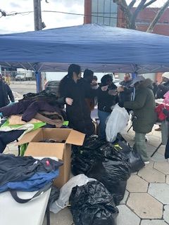 People receiving clothing under a blue canopy outside. Clothes are on tables and in bags.