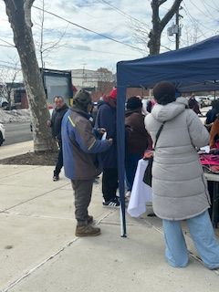 People line up at a blue tent outdoors. One person wears a light gray coat and blue jeans.