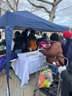 People at a table under a blue tent; items are on the table and in shopping carts; outdoors.
