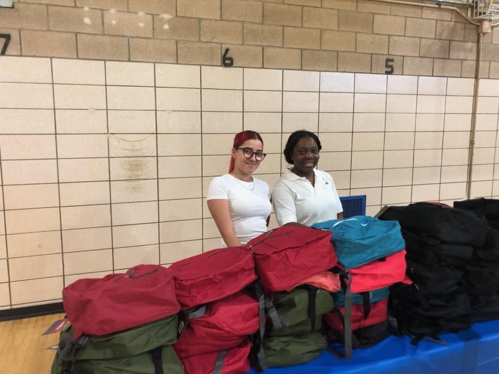 Two women are standing next to a table full of backpacks.