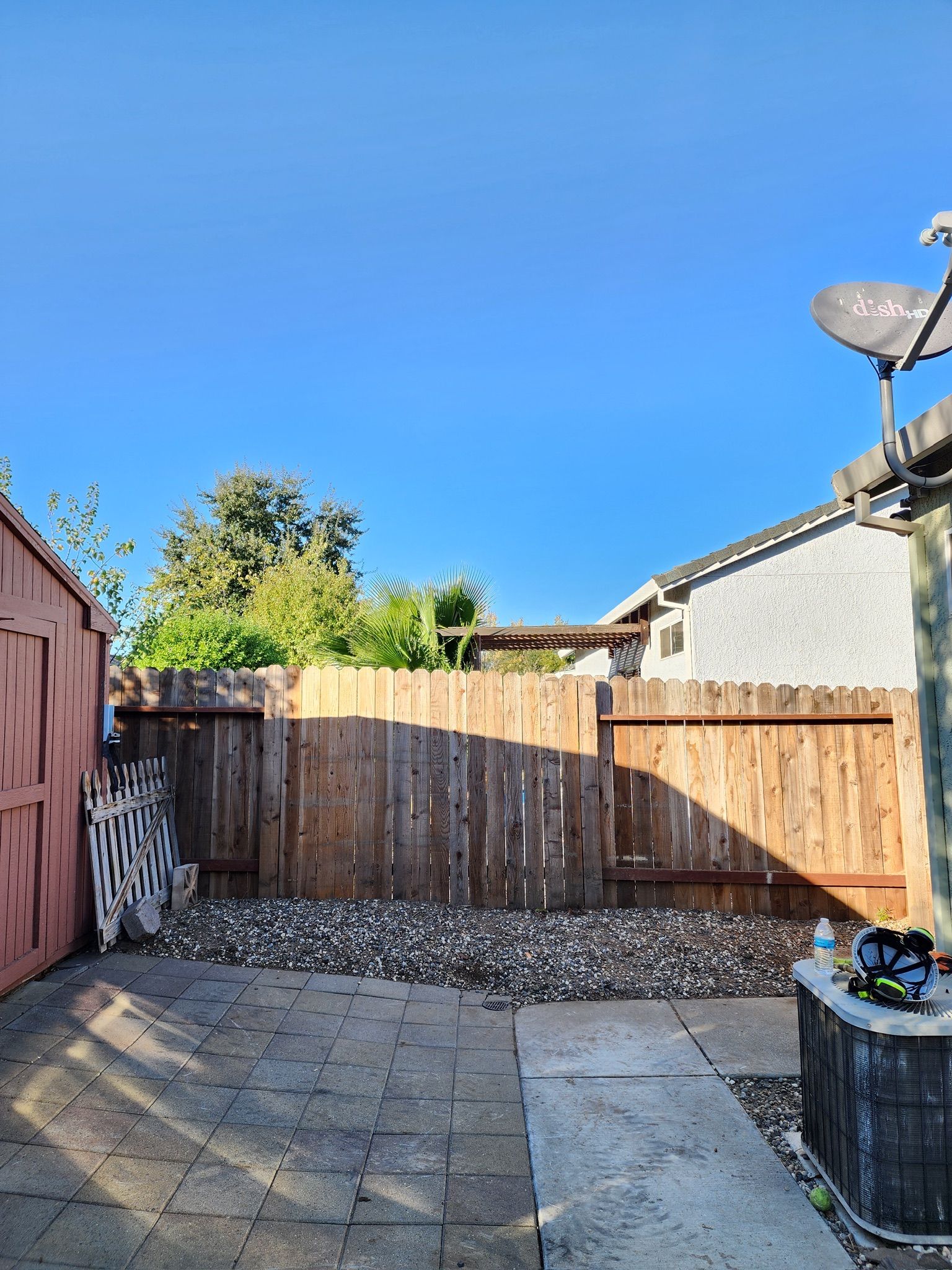 Backyard with wooden fence, shed, and patio under a bright blue sky.