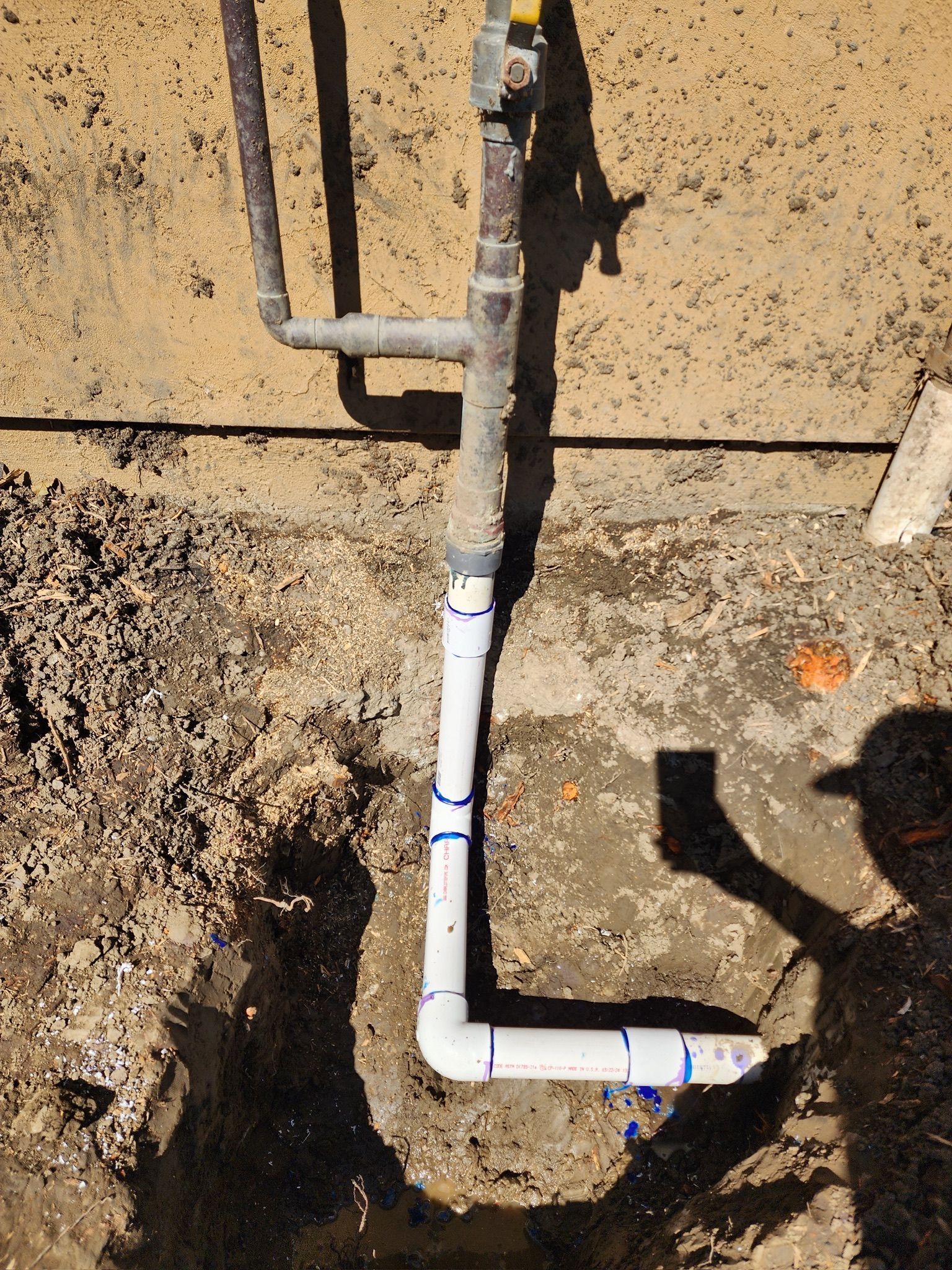 Pipes in a trench, connected with white fittings, against a tan wall.