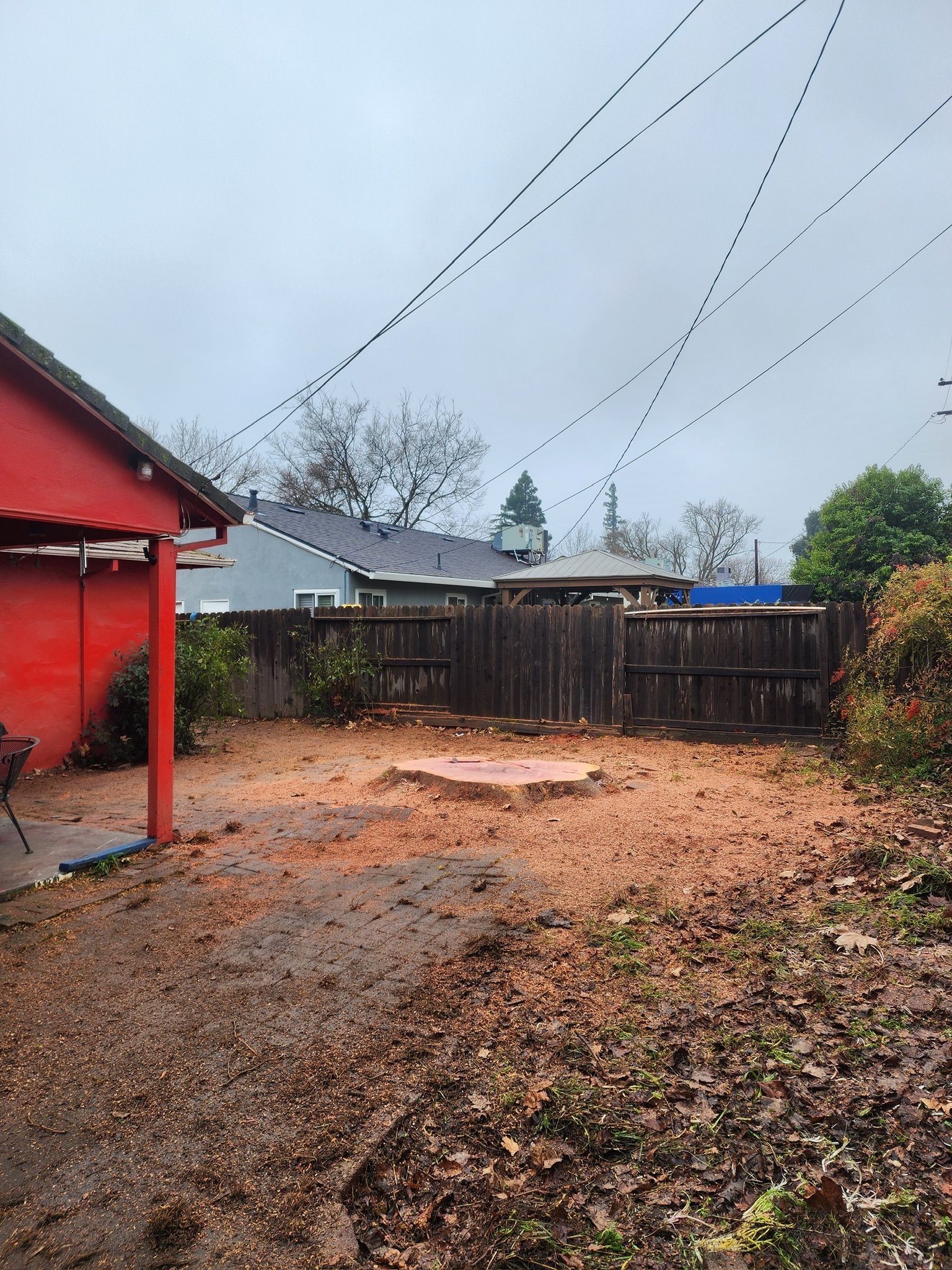 Backyard with red building, wooden fence, tree stump, power lines overhead, overcast sky.