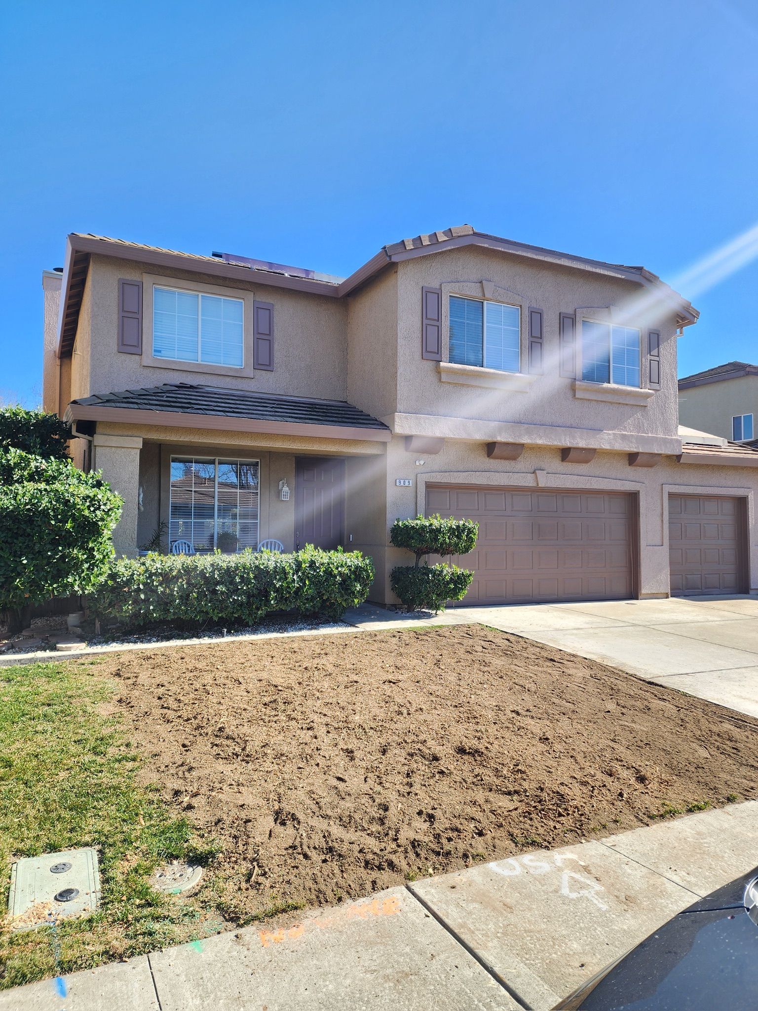 Two-story beige house with brown shutters, a three-car garage, and dry lawn under a clear, blue sky.