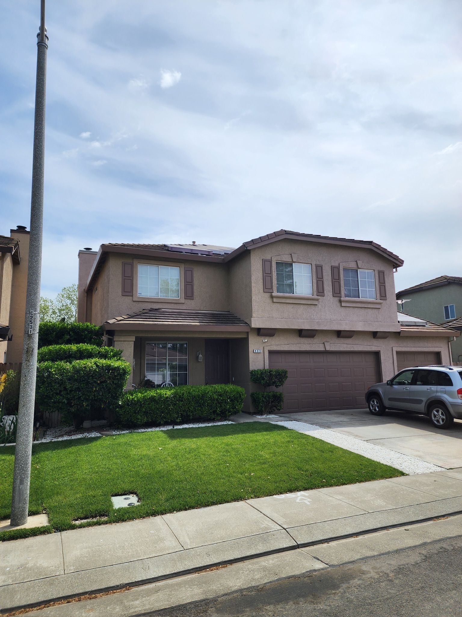 Two-story beige house with a green lawn, shrubbery, and a car parked in the driveway.