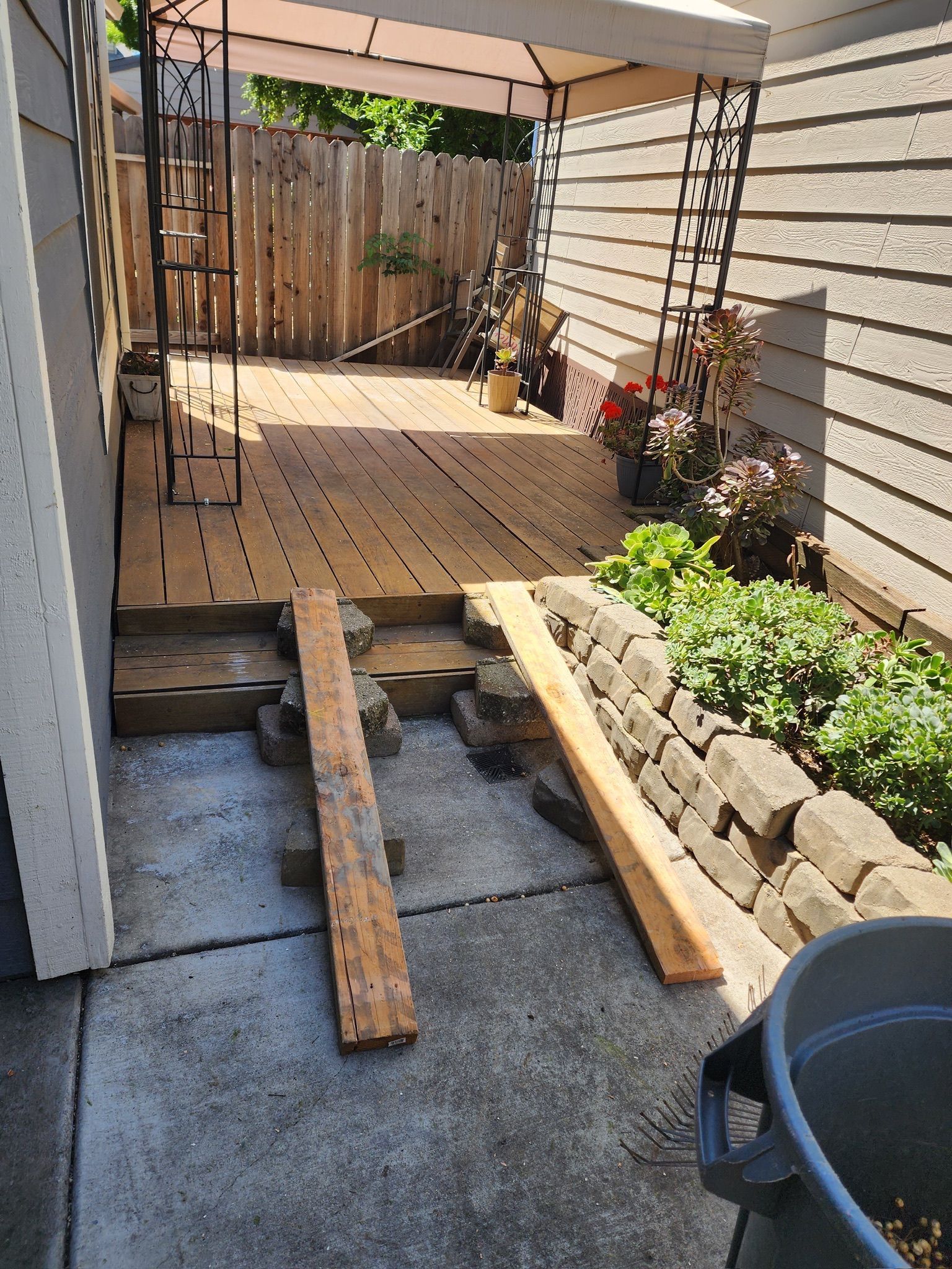 A small deck with ramp under construction, next to a garden bed and concrete patio.