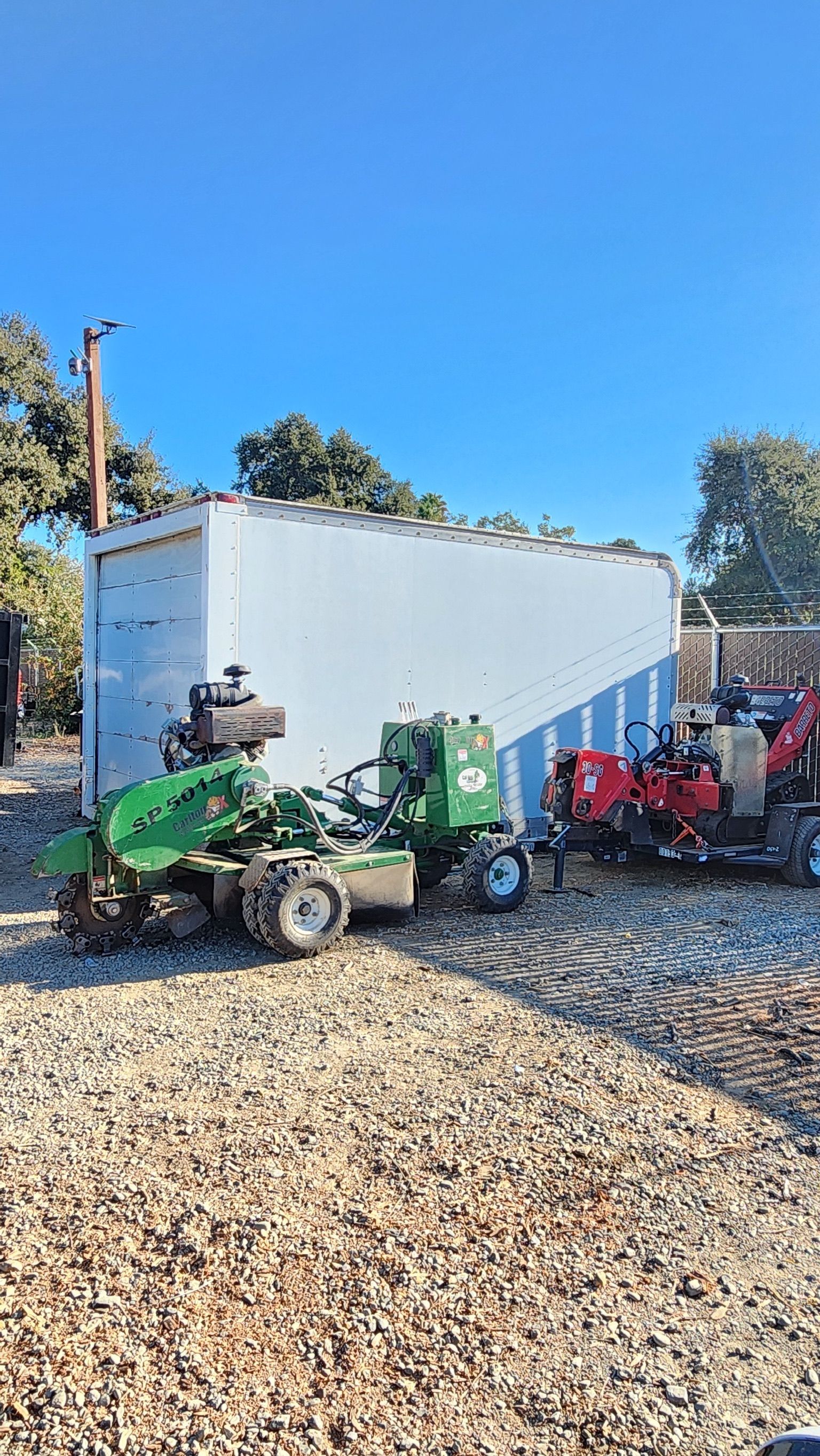 Green stump grinder next to a white building; gravel ground, blue sky.