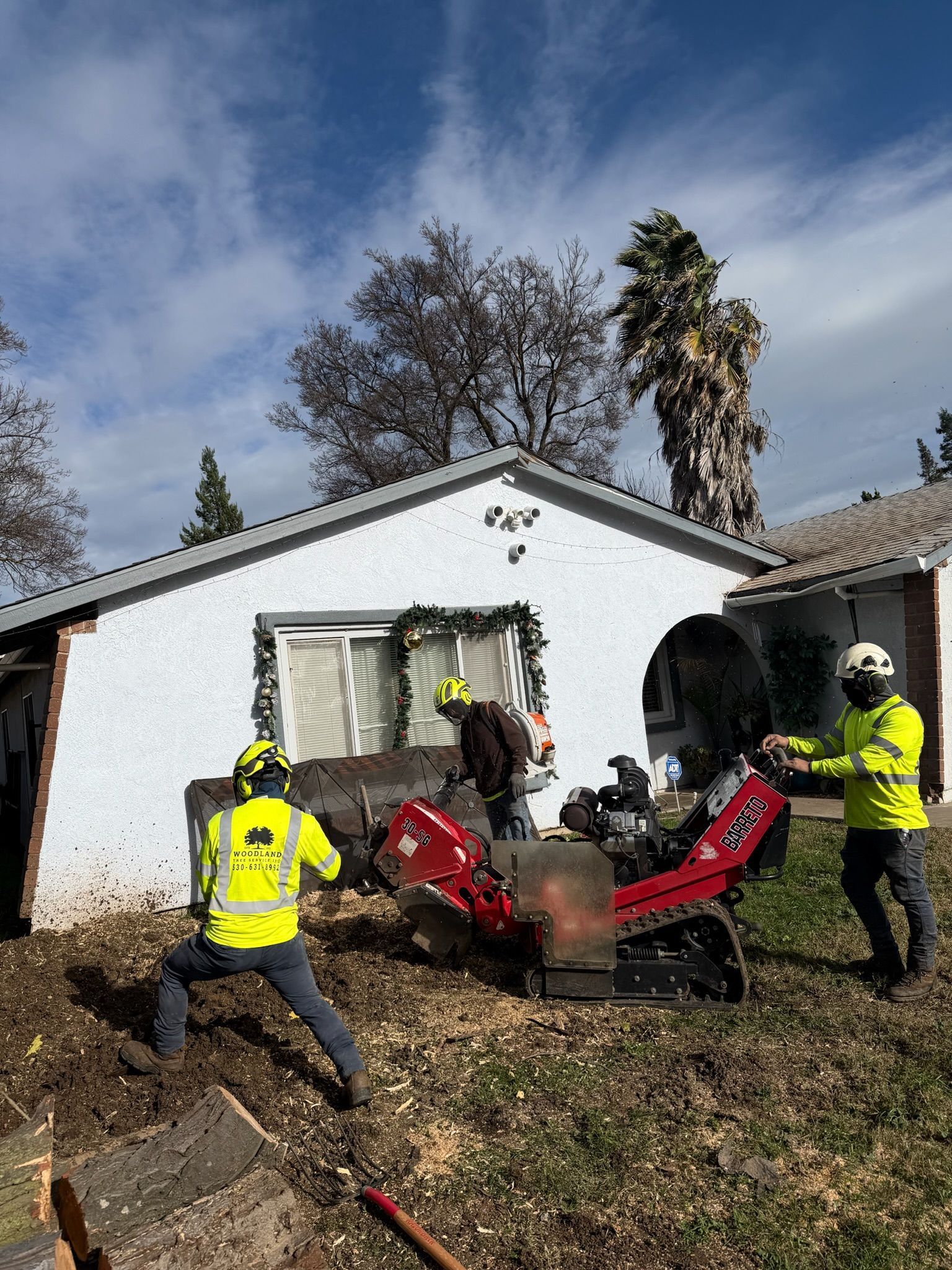 Three workers in safety vests operating a wood chipper near a white house with a cloudy sky.