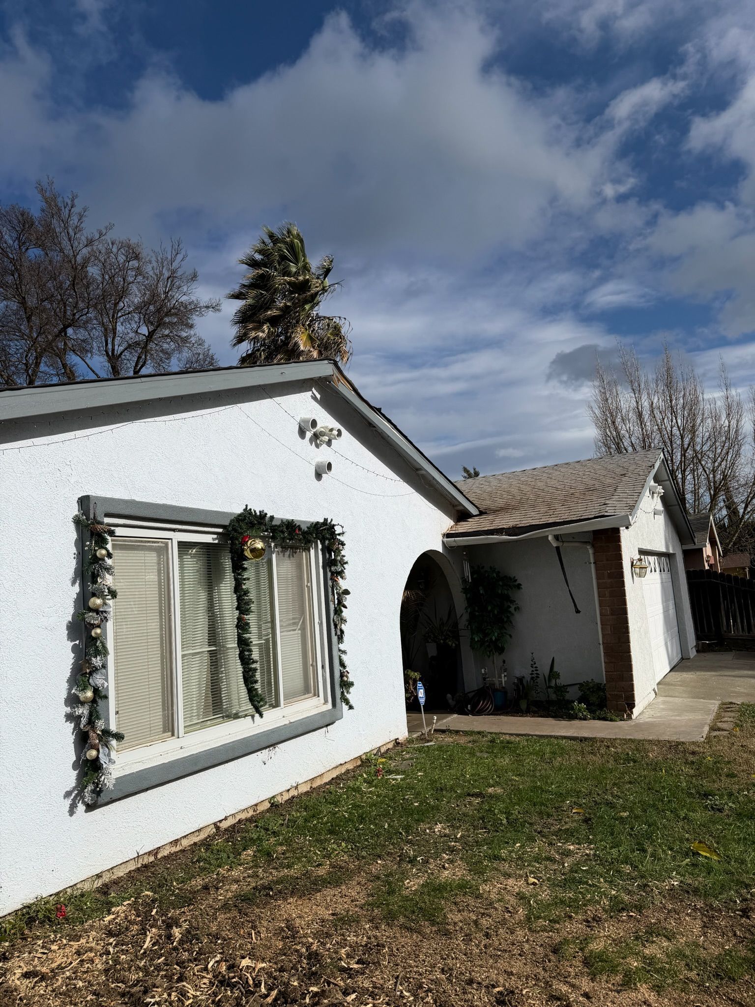 White house with gray trim, Christmas garland on a window, under a cloudy blue sky.