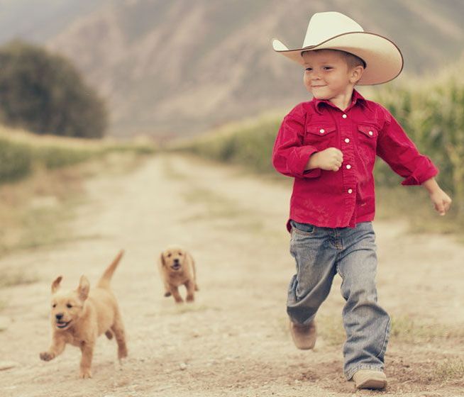 Boy in cowboy hat, red shirt, and jeans runs down dirt road with two golden retriever puppies.