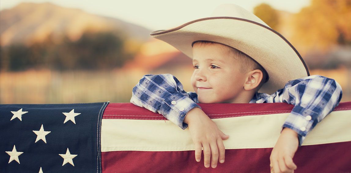 A young child wearing a cowboy hat leans on an American flag, smiling outdoors.