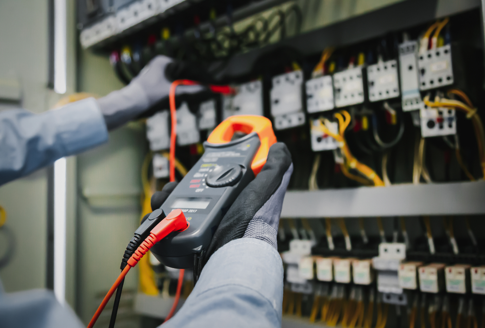 A person in work gloves uses a digital clamp meter to test electrical components inside a control panel.