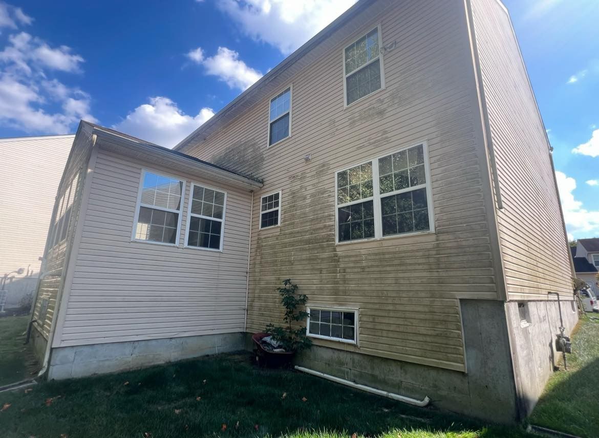 Tan house exterior with stained siding, several windows, and a green lawn under a partly cloudy blue sky.