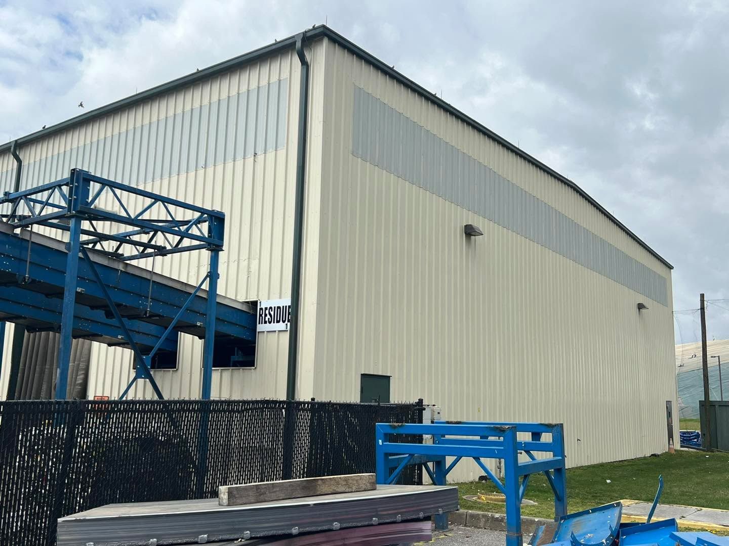 Beige industrial building with a blue conveyor system and black fence.